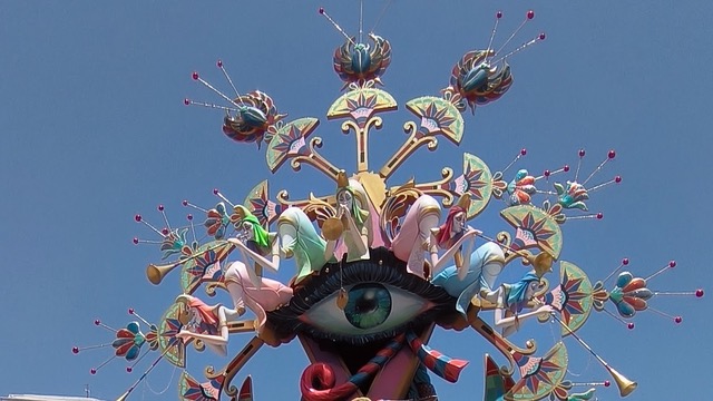 Een kleurrijke en gedetailleerde sculptuur van het Las Hogueras Festival. De sculptuur toont een groot oog in het midden, omringd door diverse abstracte en fantasierijke figuren en patronen, waaronder bloemen en krullende vormen, allemaal tegen een heldere blauwe hemel.